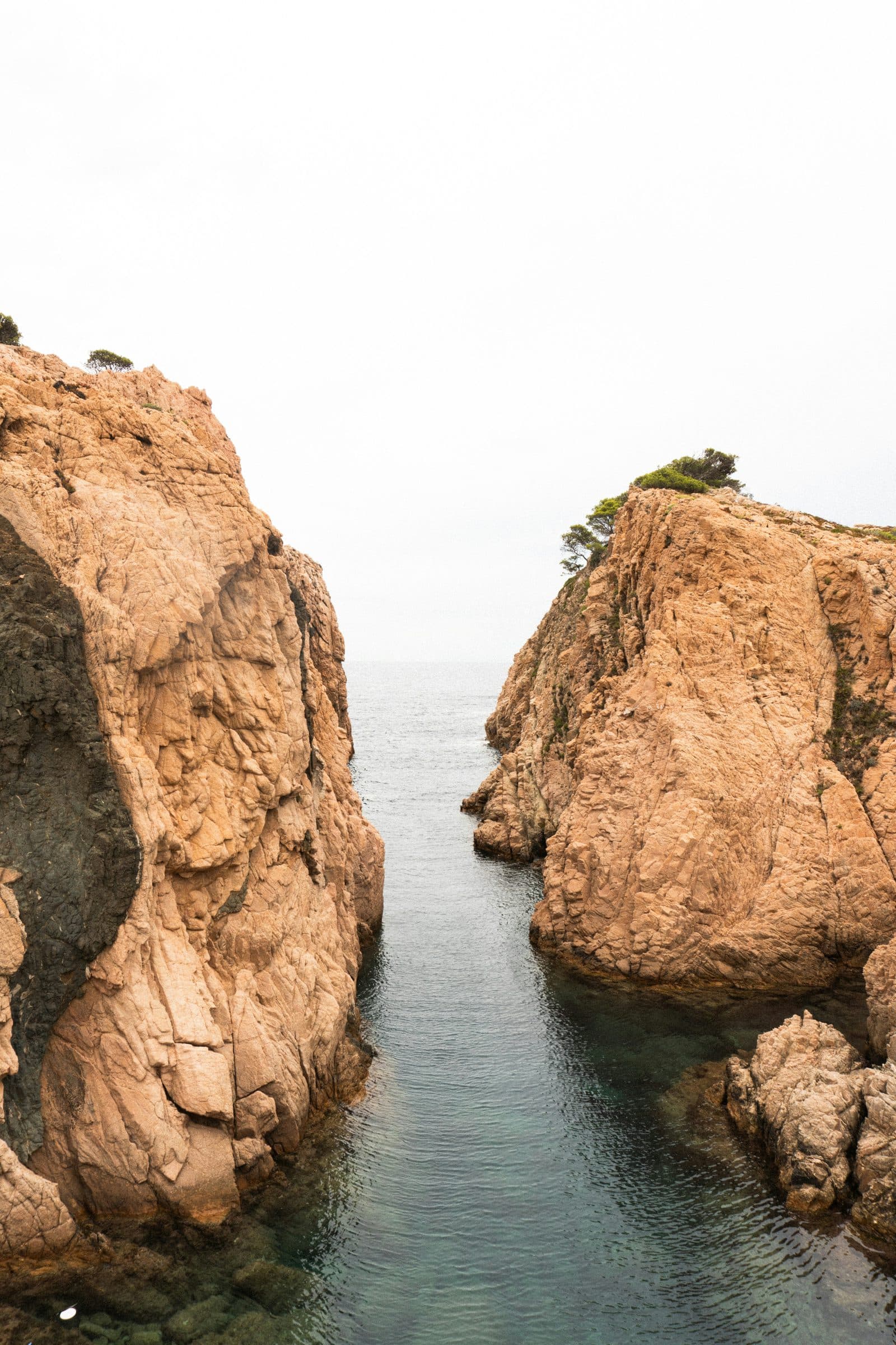 Un estrecho paso de agua entre dos paredes de roca rojiza en una cala de Begur, con pinos en lo alto de los acantilados