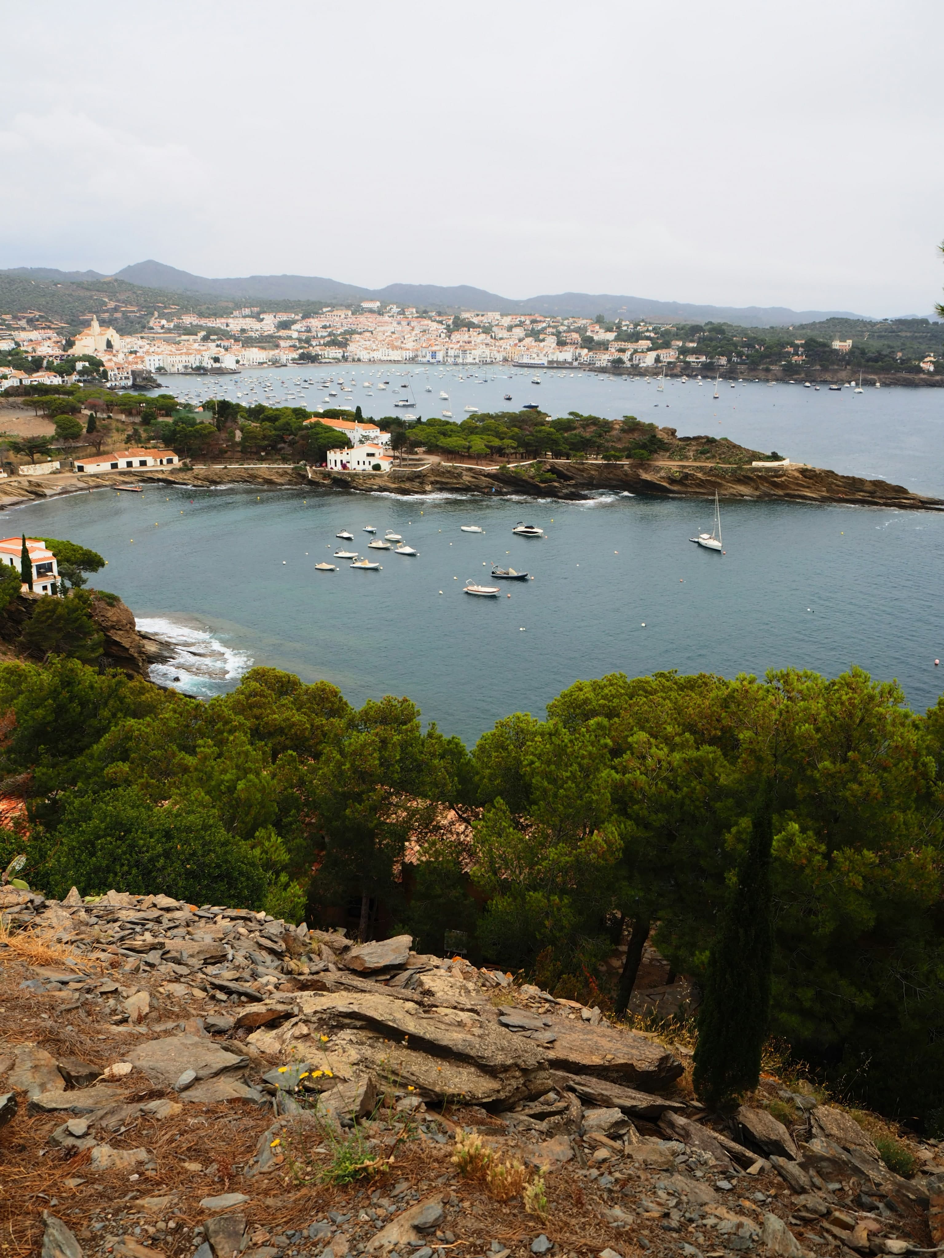 Una cala recogida cerca de Cadaqués con barcas fondeadas y pinos, y el pueblo blanco al fondo bajo un cielo cubierto