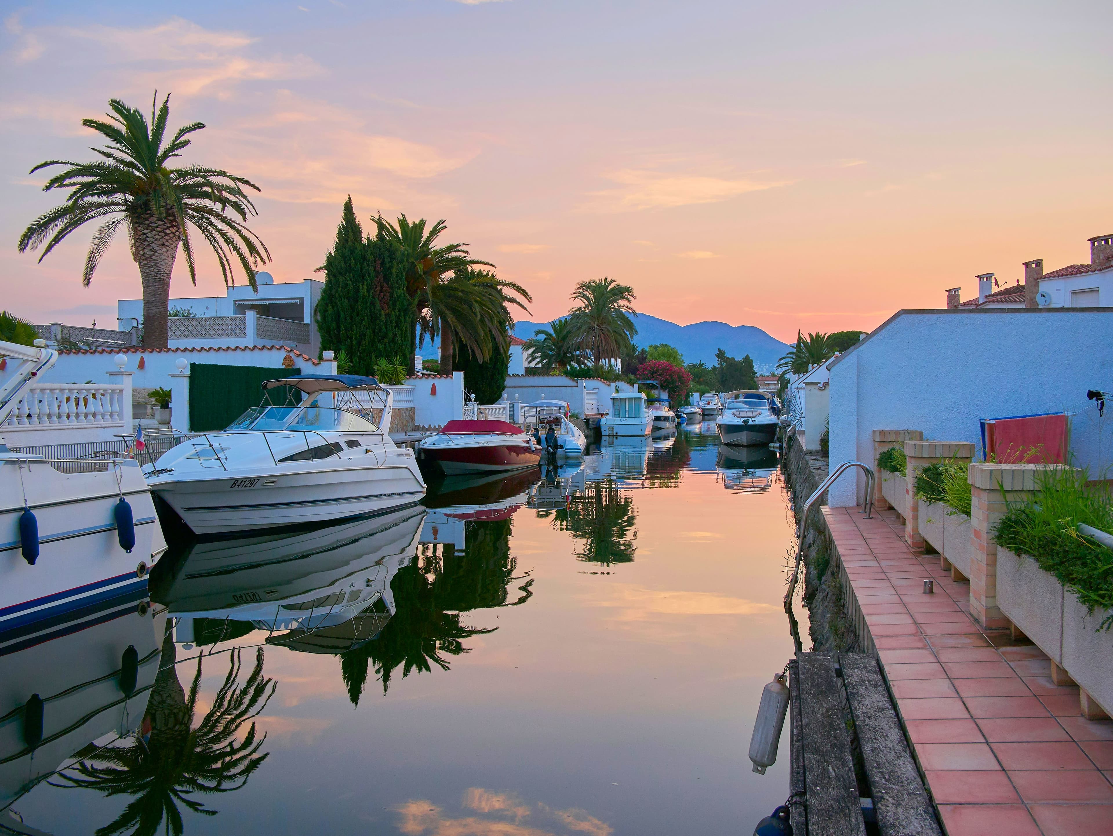 Un canal de Empuriabrava al atardecer, con barcas amarradas a la puerta de las casas, palmeras y el cielo rosado reflejado en el agua quieta
