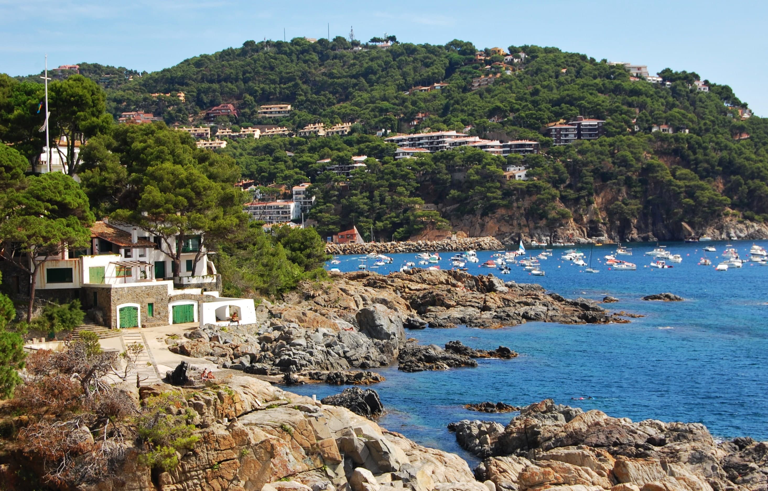 La costa rocosa del camí de ronda de Llafranc, con una antigua casa de pescadores de puertas verdes y barcas fondeadas en la bahía