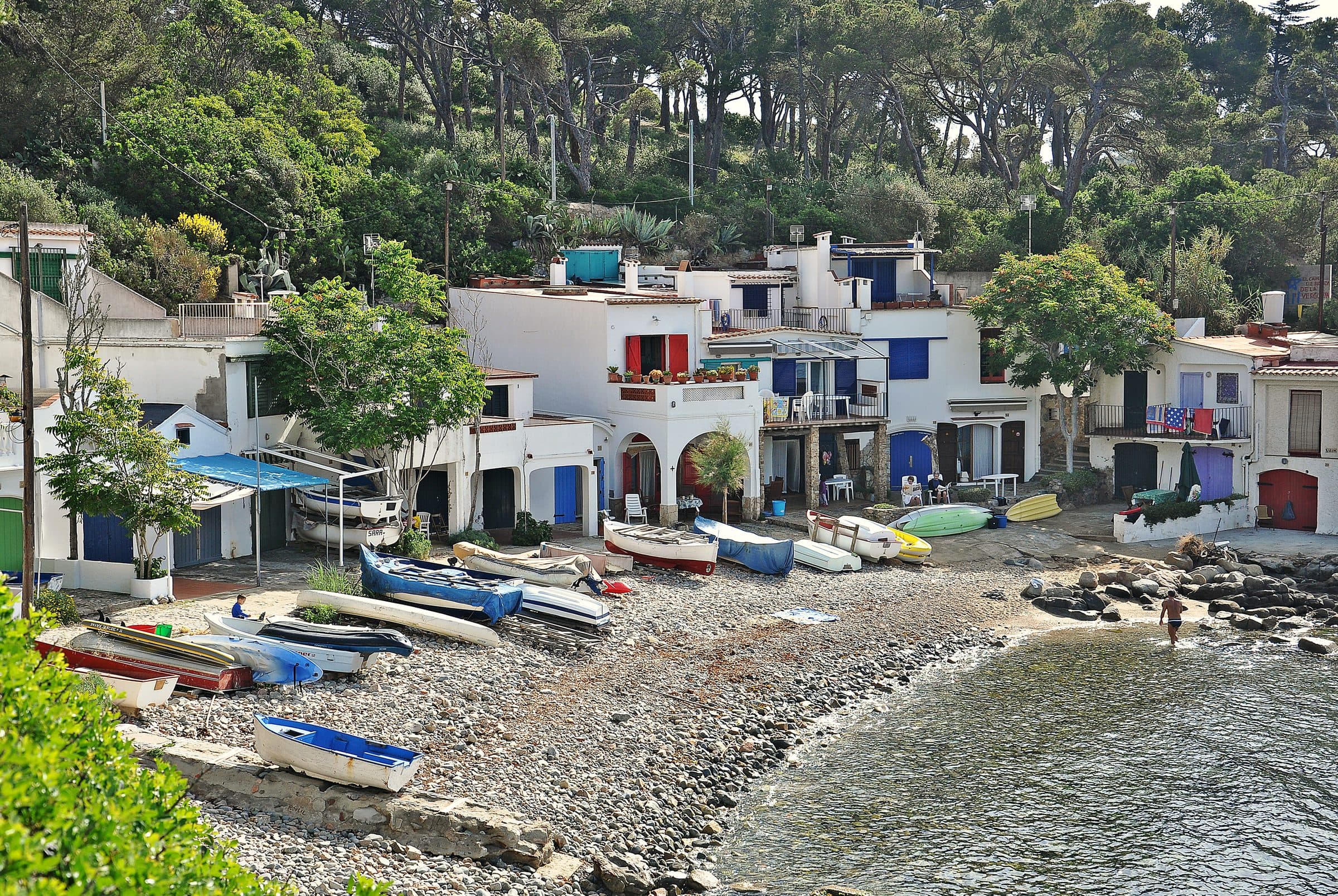 Casetas de pescadores blancas y ocres de Cala S'Alguer, apoyadas sobre la roca junto a barcas varadas en la orilla
