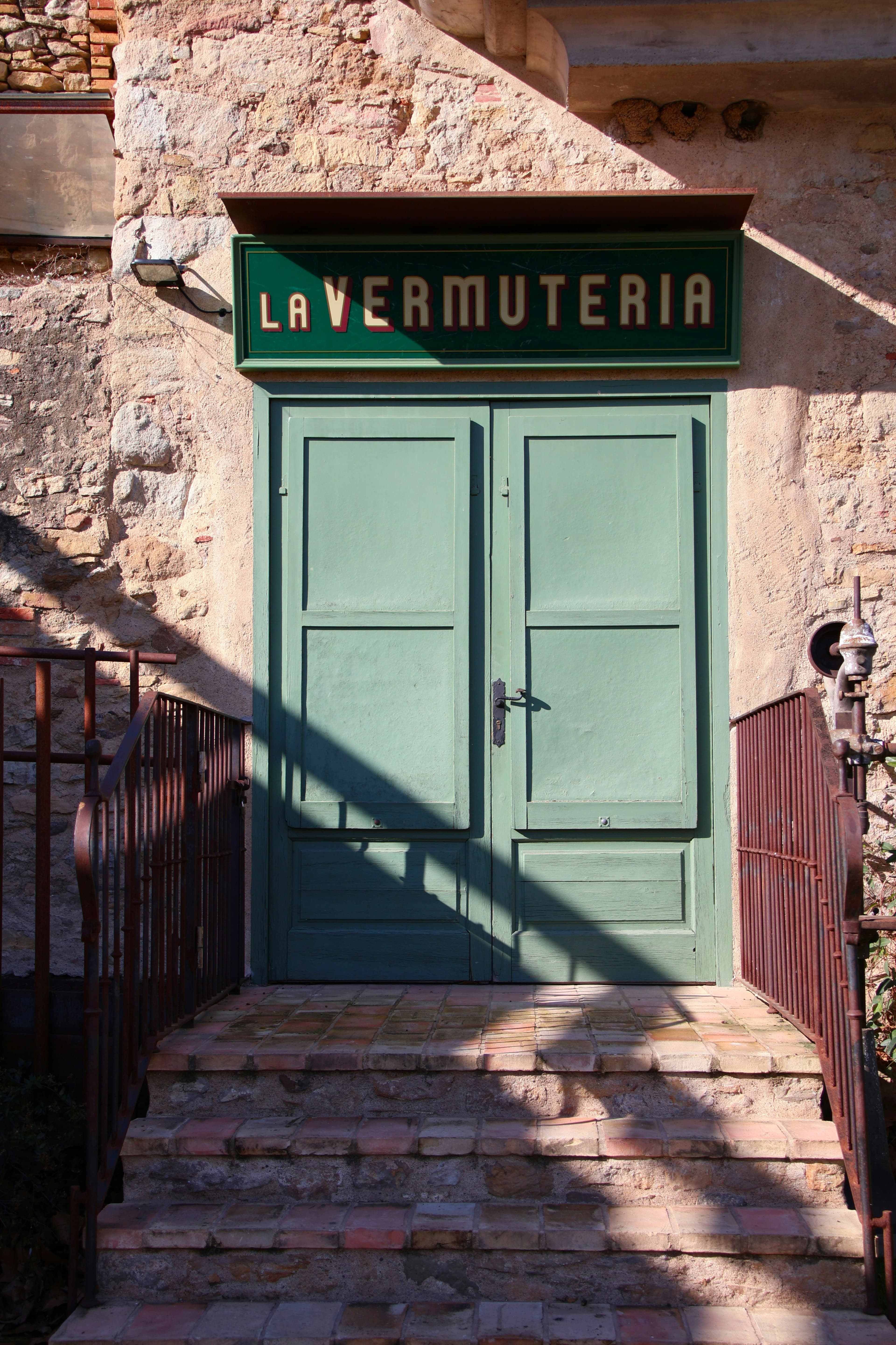 La puerta verde de una vermutería en una calle de Peratallada, bajo un cartel pintado a mano y sobre unos escalones de ladrillo