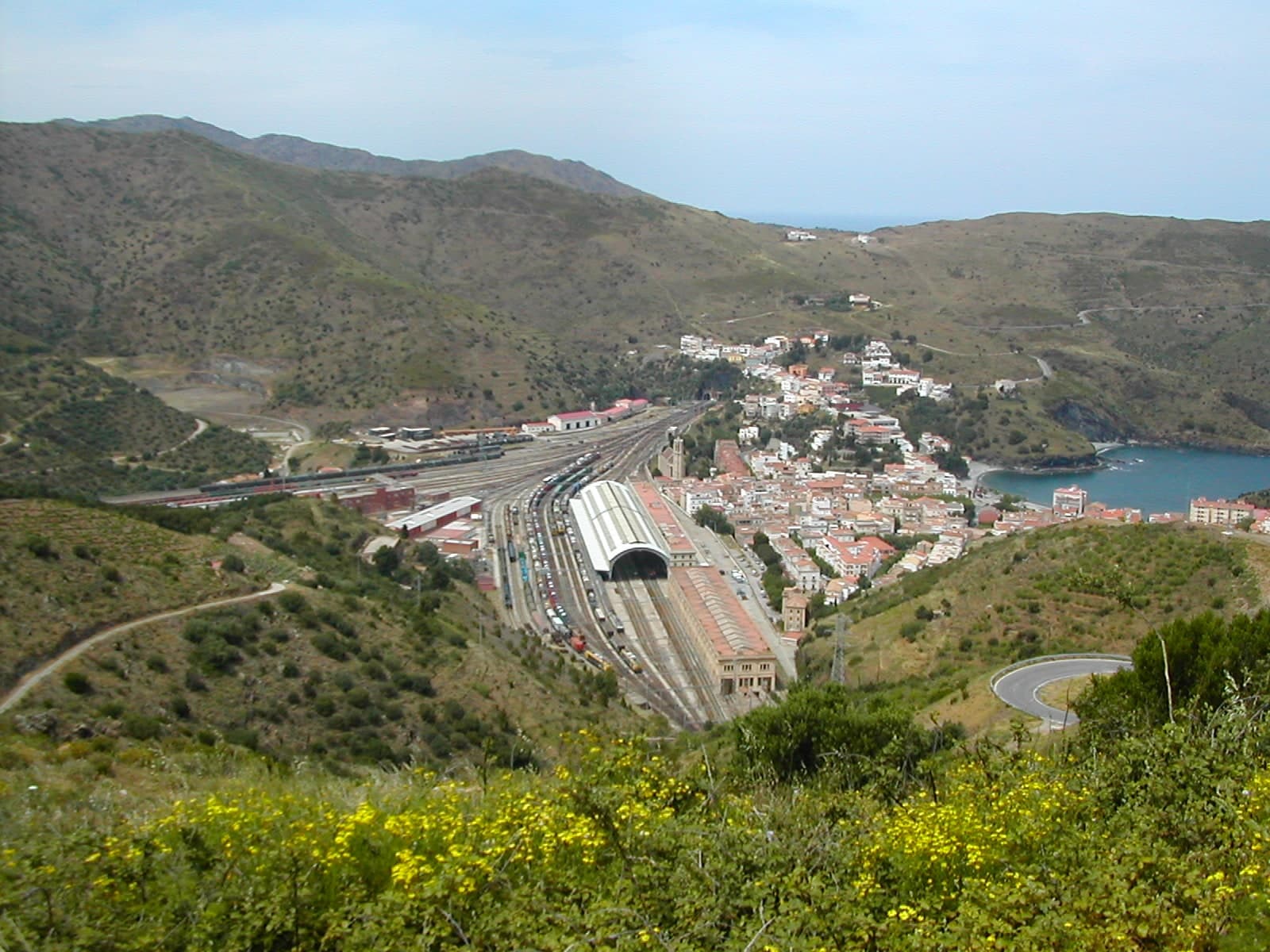 Vista aérea de Portbou con su gran estación de tren fronteriza y los andenes cubiertos, encajada entre montañas junto a la bahía
