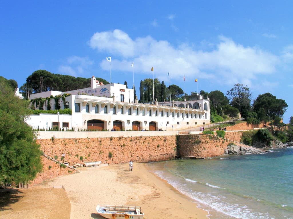 La fachada blanca de arcos del Hostal La Gavina sobre la playa de S’Agaró, con banderas ondeando y una barca en la arena
