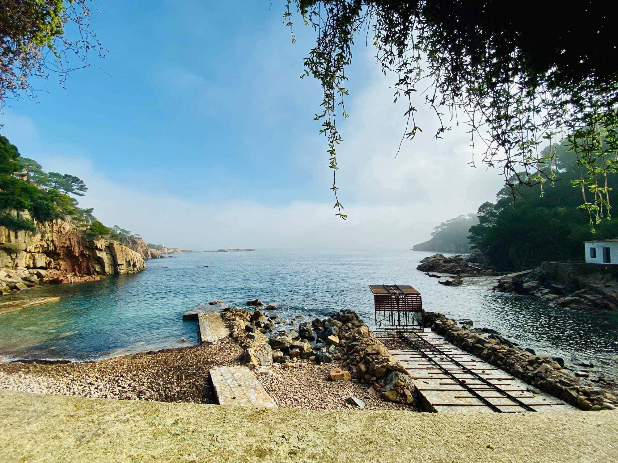 Una cala de Begur de aguas turquesas y transparentes, con un pequeño embarcadero de piedra y pinos sobre la orilla