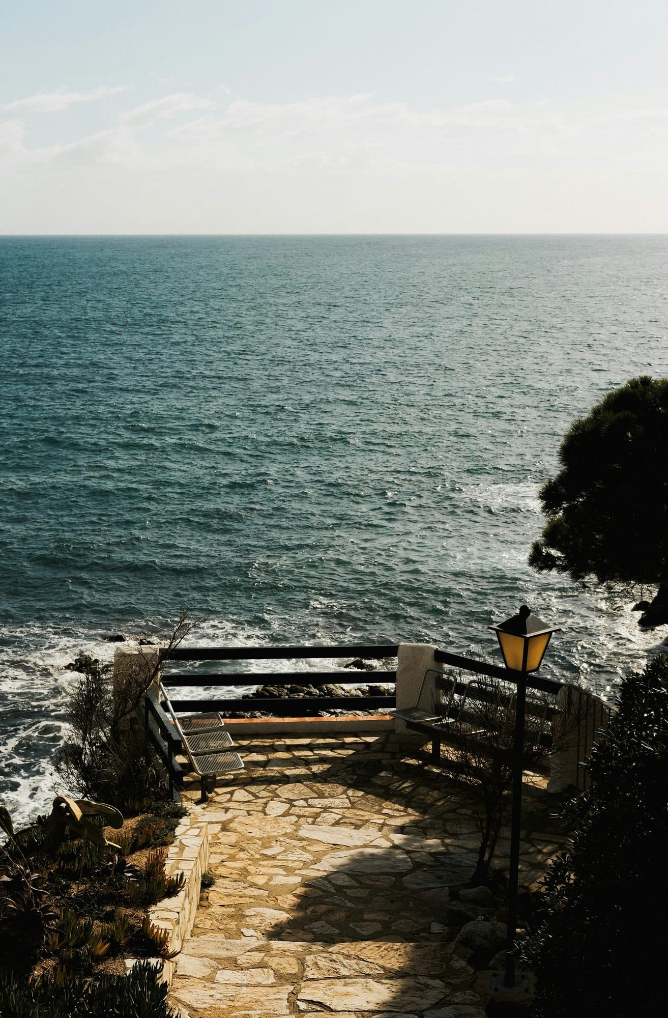 Un mirador de piedra sobre el mar en el Camí de Ronda, con bancos, una farola y pinos alrededor