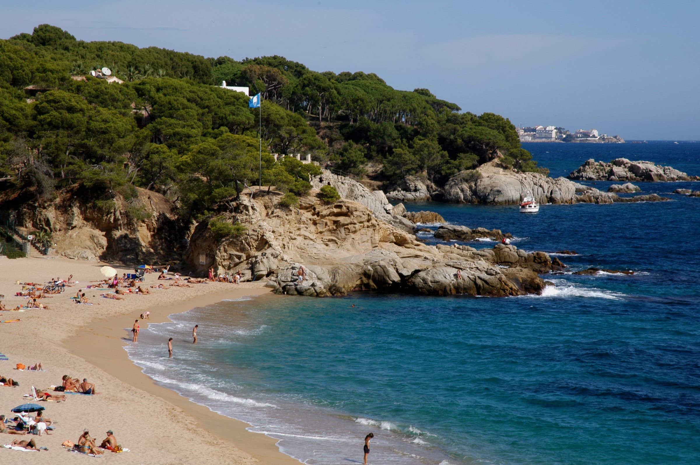 Cala Rovira, una playa de arena dorada entre roca clara y pinos, con bañistas y un velero fondeado