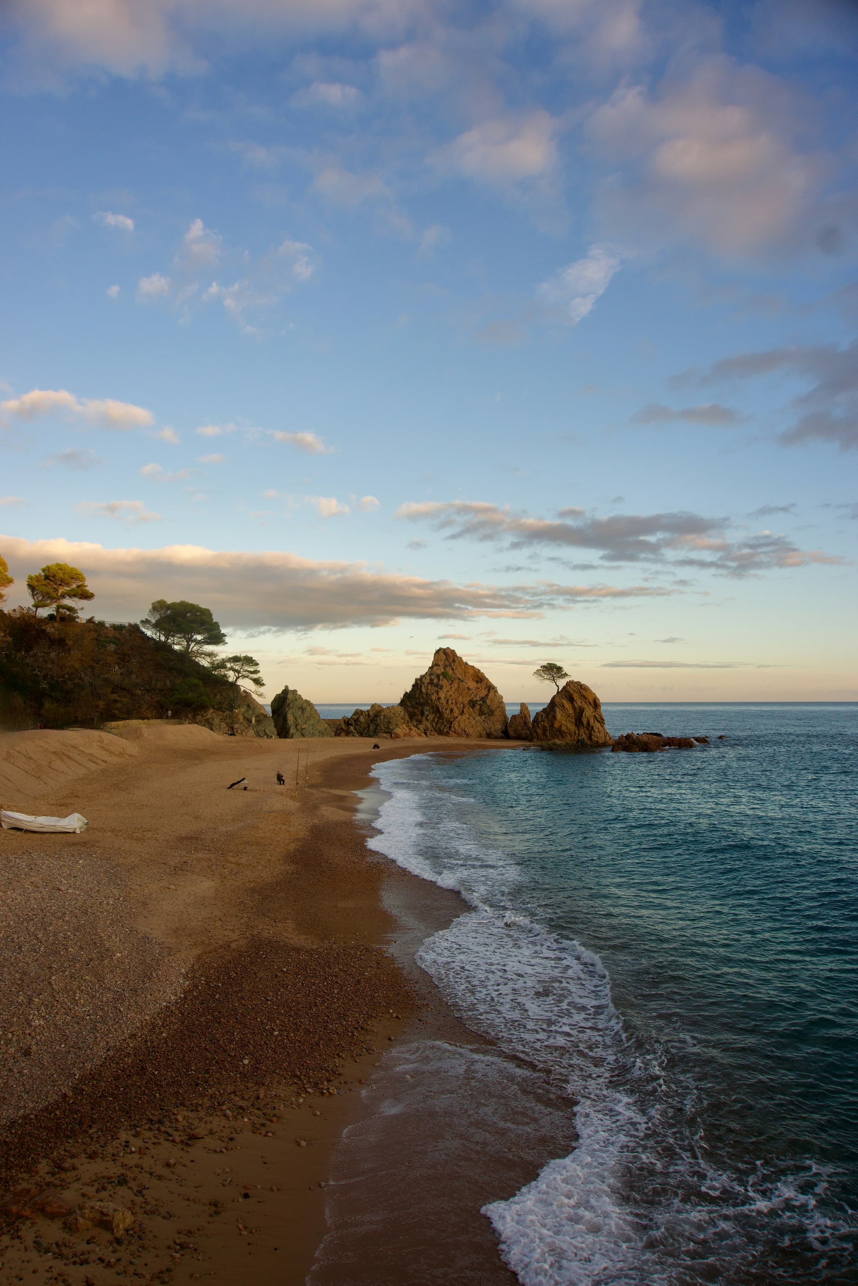 Una playa de Tossa de Mar al atardecer, con las rocas puntiagudas y un pino solitario recortados sobre el mar